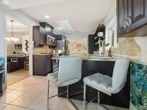 Una cocina moderna con armarios oscuros, una isla central con taburetes y un área de comedor con vista al salón.
