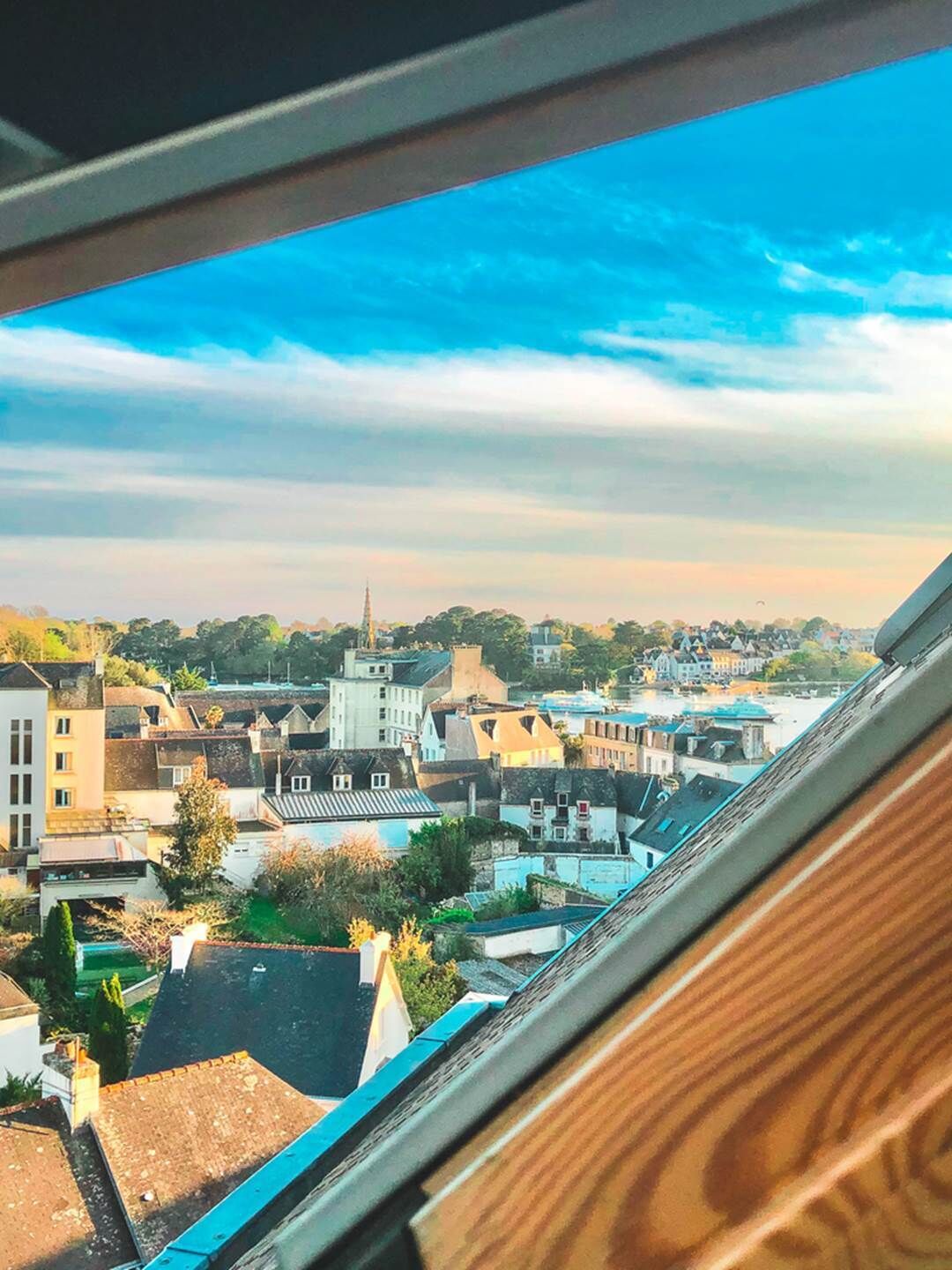 Blick aus einem Fenster auf eine Stadt mit verschiedenen Gebäuden, darunter ein Kirchturm, und einem klaren Himmel.