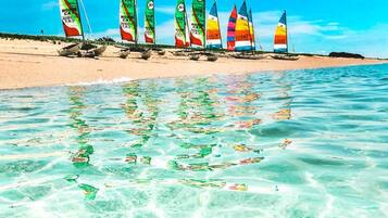 Uma praia com águas calmas e transparentes e vários veleiros com velas coloridas.