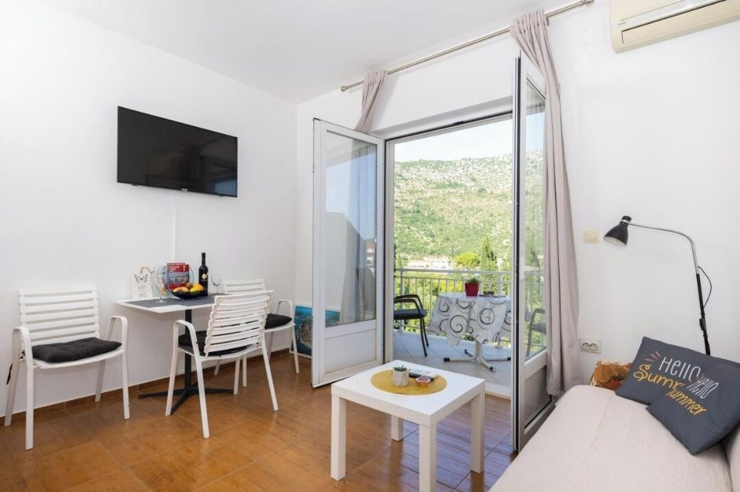 Une pièce avec une télévision, une table avec des chaises, une petite table avec une plante, un canapé avec un coussin et un balcon avec vue.
