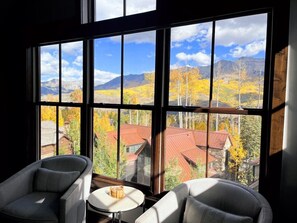 A room with large windows offering a view of a mountainous landscape with autumn foliage. There are two chairs and a small round table in the room. - The Veranda at Trails Edge - penthouse views! (Mountain Village)