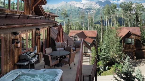A wooden cabin with a patio, outdoor grill, and hot tub, overlooking a mountainous landscape.