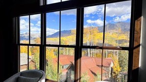 A room with large windows offering a view of autumn foliage and mountains. There are two chairs and a small table.