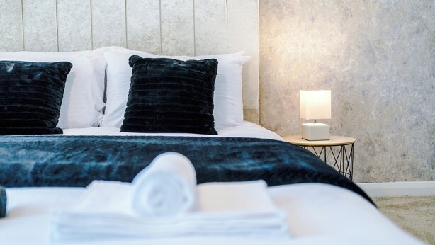 Un lit bien fait, avec des oreillers de couleurs foncées et claires, une lampe de chevet blanche et une serviette pliée posée sur le lit.