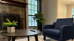 A living room with a stone fireplace, a round coffee table, a blue armchair, and a large window with a view.
