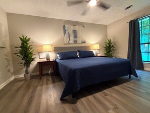 Une chambre à coucher comprenant un lit, deux tables de chevet, deux lampes, deux plantes en pot, un ventilateur de plafond et une fenêtre avec des rideaux.
