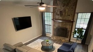 A living room with a stone fireplace, a ceiling fan, a flat-screen TV, a gray sofa, a blue armchair, and a coffee table.