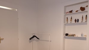 Une pièce avec un plancher en bois, une table blanche recouverte d’une nappe à motifs, une chaise rouge, un portant à vêtements et des étagères ornées d’objets décoratifs.