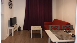 Une petite pièce bien éclairée, avec un canapé rouge, une table blanche ornée d’un chemin de table à motifs, une petite table blanche, un téléviseur posé sur un meuble blanc et une grande fenêtre avec un rideau foncé.