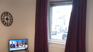 Une chambre d’hôtel avec un lit, un téléviseur posé sur un meuble en bois, une horloge au mur et une fenêtre avec des rideaux.