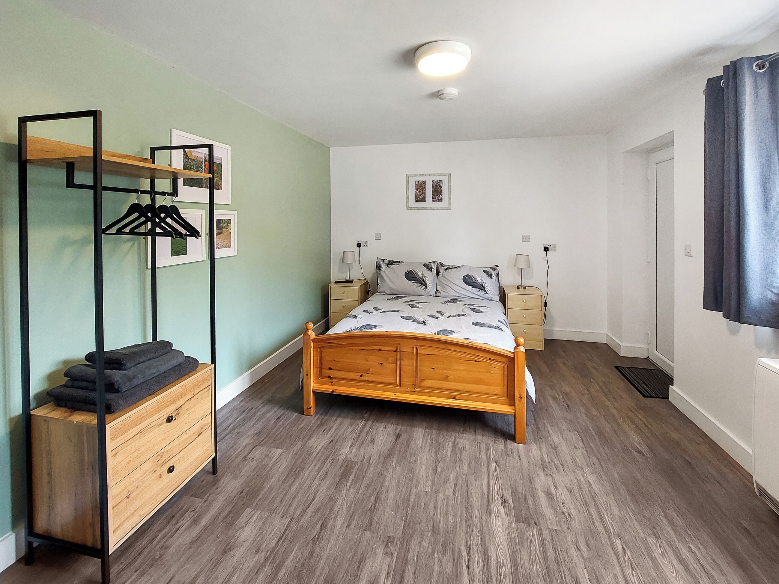 A bedroom with a wooden bed, a bedside table with a lamp, a dresser, a hanging clothes rack, and a ceiling light.