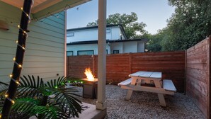 A patio area with a wooden fence, a concrete planter with a plant, a white picnic table, and a fire feature. - Upstay Entertainment Hub Games & Firepit (Austin)
