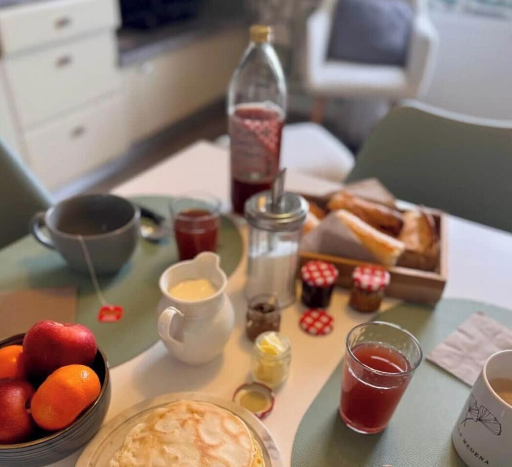 A breakfast setting with a plate of pancakes, a bowl of fruit, a bottle of juice, a cup of coffee, and a small jar of jam.