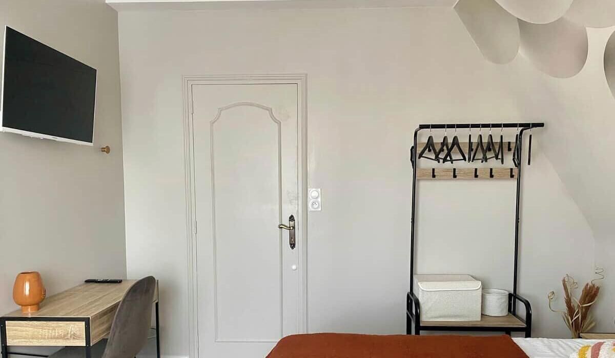Quarto com uma cama, uma escrivaninha com cadeira, uma TV fixada na parede e um cabideiro com cadeira.