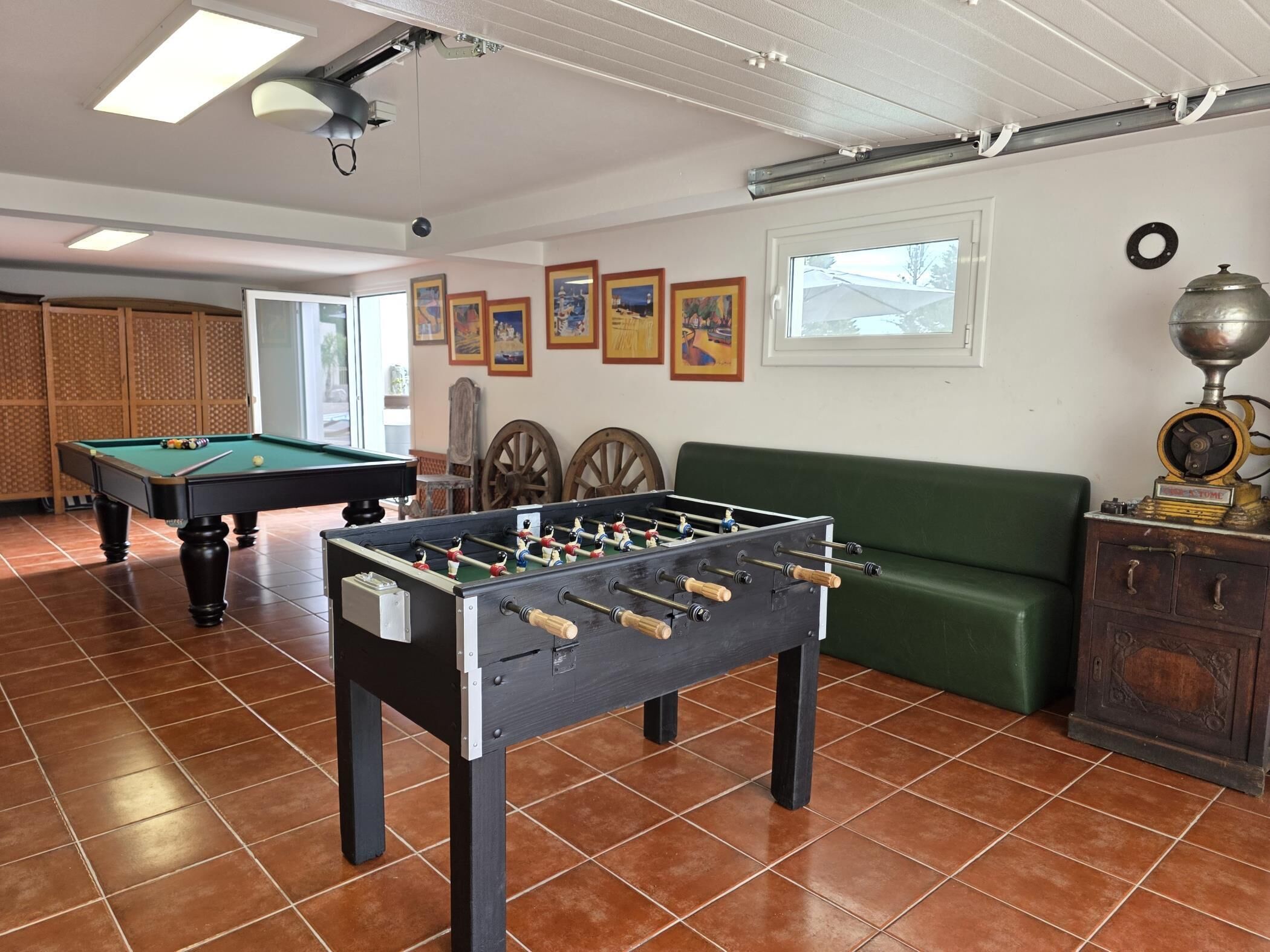 Una habitación con mesa de fútbol, mesa de billar y sofá. Hay cuadros en la pared y una ventana con vistas a árboles.