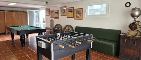 Una habitación con mesa de fútbol, mesa de billar y sofá. Hay cuadros en la pared y una ventana con vistas a árboles.