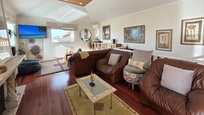 A spacious living room with a wooden ceiling, a large TV, and a view of the sea through a window.