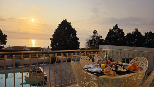 A dining table with chairs on a balcony overlooking a swimming pool and the sunset.