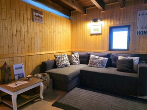 Une pièce chaleureuse avec un mur en bois, un canapé gris agrémenté de coussins à motifs, une petite table avec une lampe et une fenêtre donnant sur un beau paysage.