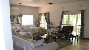 Ein Wohnzimmer mit einer Couch, einem Sessel und einem Couchtisch. Ein Fenster mit Vorhängen, ein Ventilator und ein kleiner Tisch mit einer Pflanze sind ebenfalls vorhanden.
