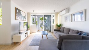 Un salon moderne avec un canapé gris, un téléviseur posé sur un meuble blanc, une petite table et une grande fenêtre donnant sur un paysage verdoyant.