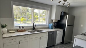 A modern kitchen with white cabinets, a stainless steel refrigerator, a double sink, and a window with blinds. - Seashell Serenity Beach Escape - Just 5 minutes from Lovely Bonita Beach Park! (Bonita Springs)