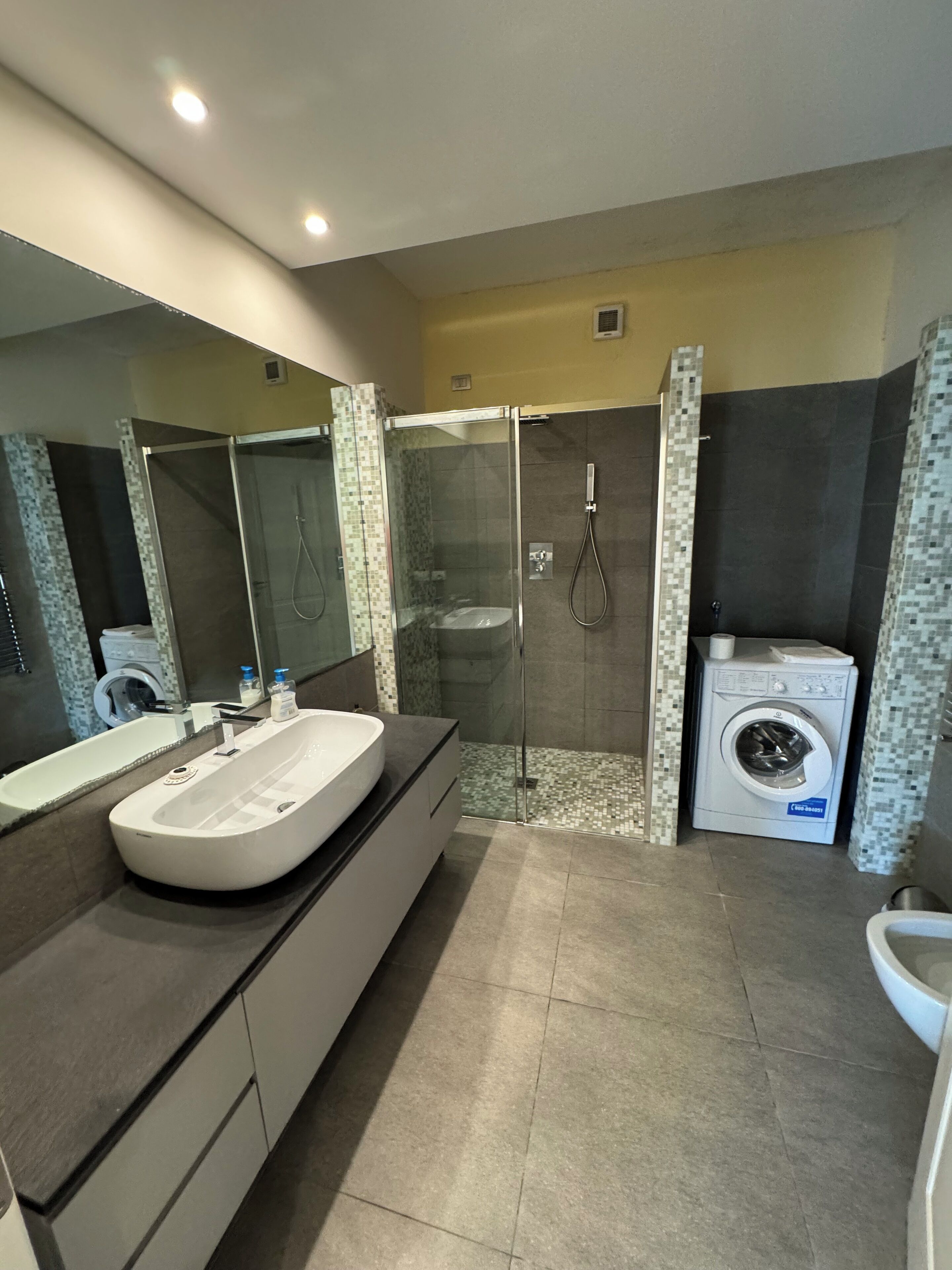 A modern bathroom with a large mirror, a freestanding bathtub, a glass-enclosed shower, and a built-in washing machine.