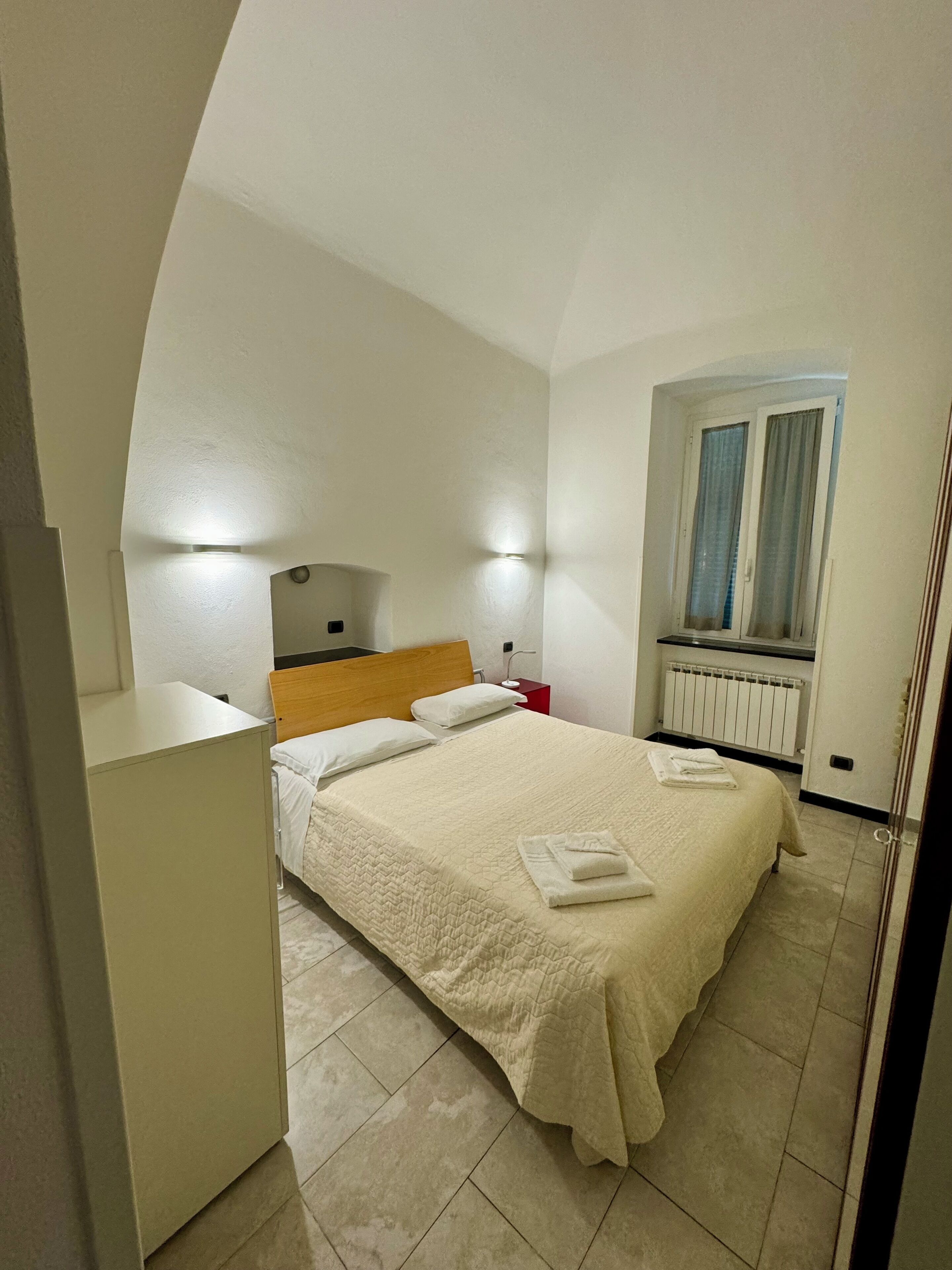 A bedroom with a bed, bedside table, lamp, window, and radiator.