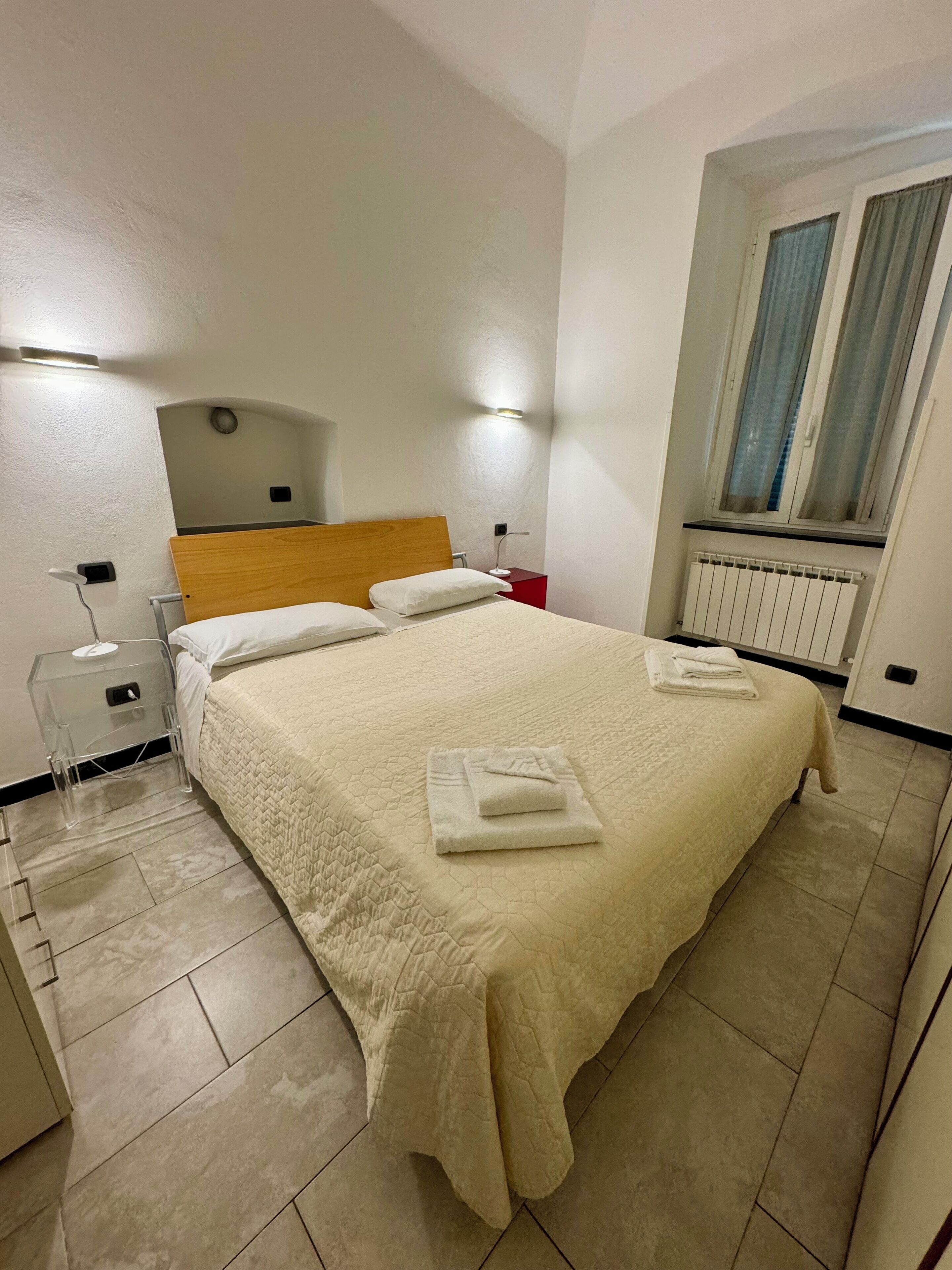 A neatly made bed with a white duvet and two pillows, a bedside table with a lamp, a red nightstand, and a window with curtains.