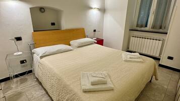 A neatly made bed with a white duvet and two pillows, a bedside table with a lamp, a red nightstand, and a window with curtains.