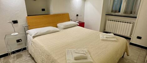 A neatly made bed with a white duvet and two pillows, a bedside table with a lamp, a red nightstand, and a window with curtains.