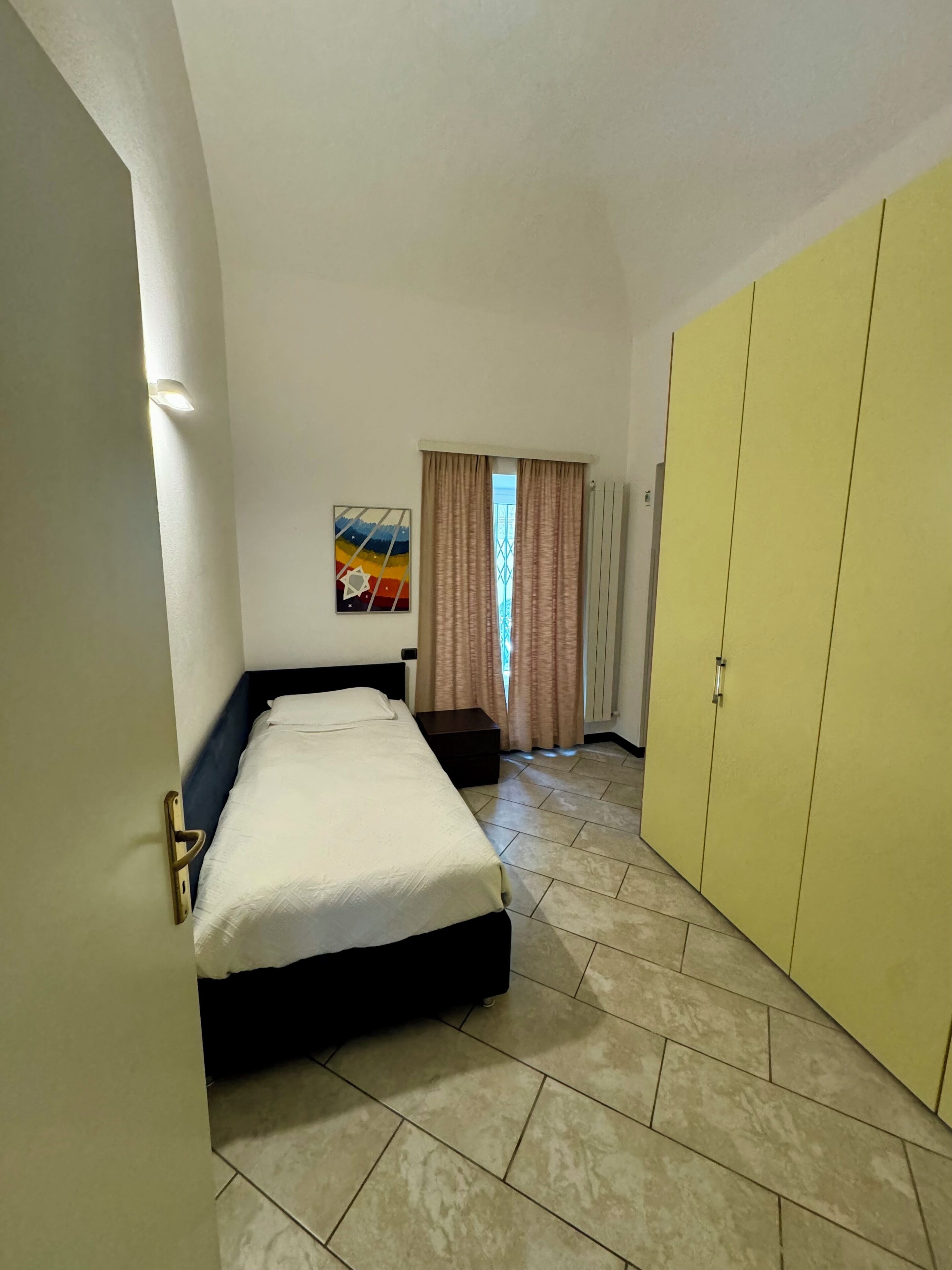 Un dormitorio con cama, armario, mesita de noche, un cuadro y una ventana con cortinas.