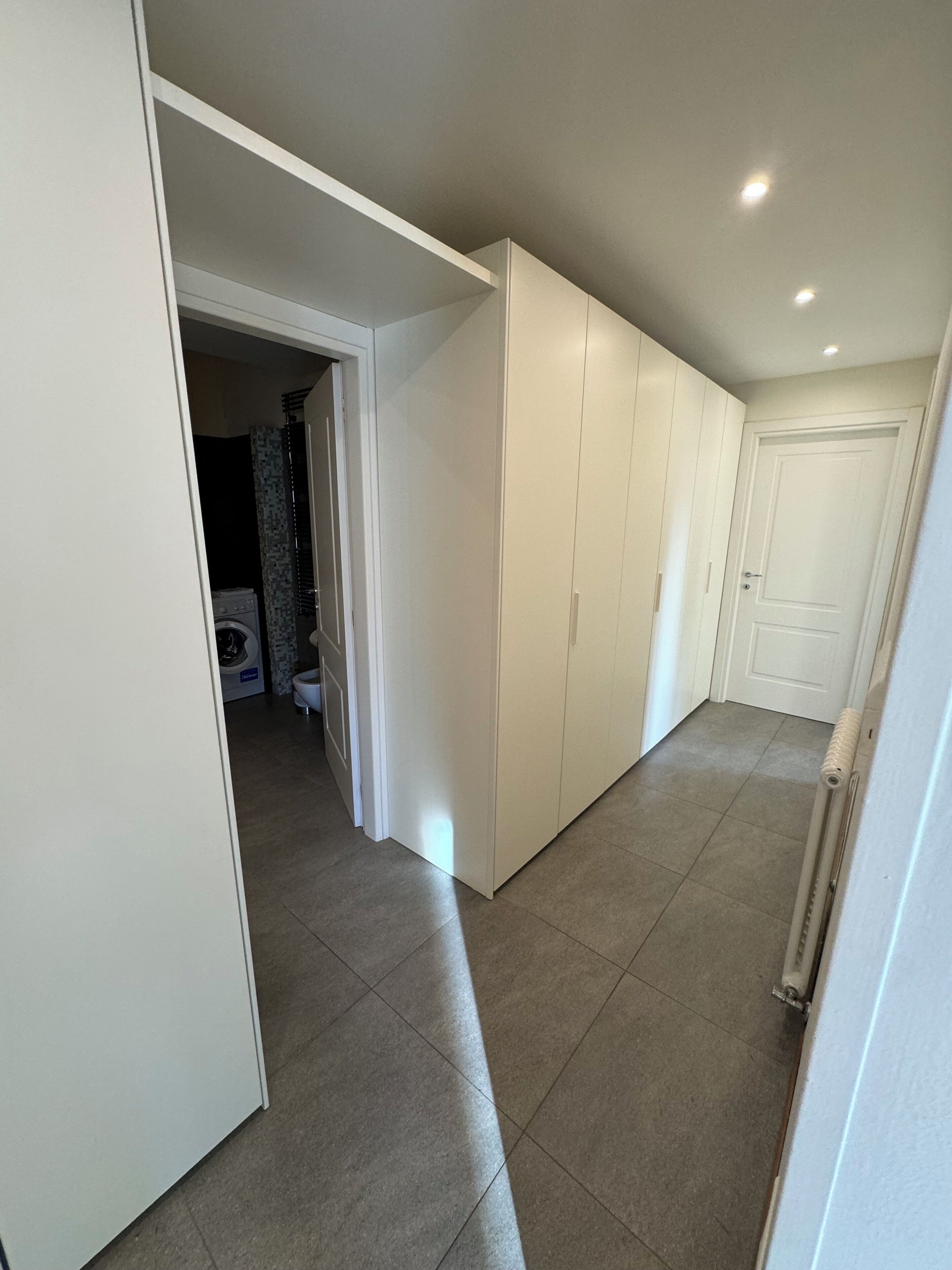 A hallway with white walls and a tiled floor, leading to a room with a washing machine and a door.