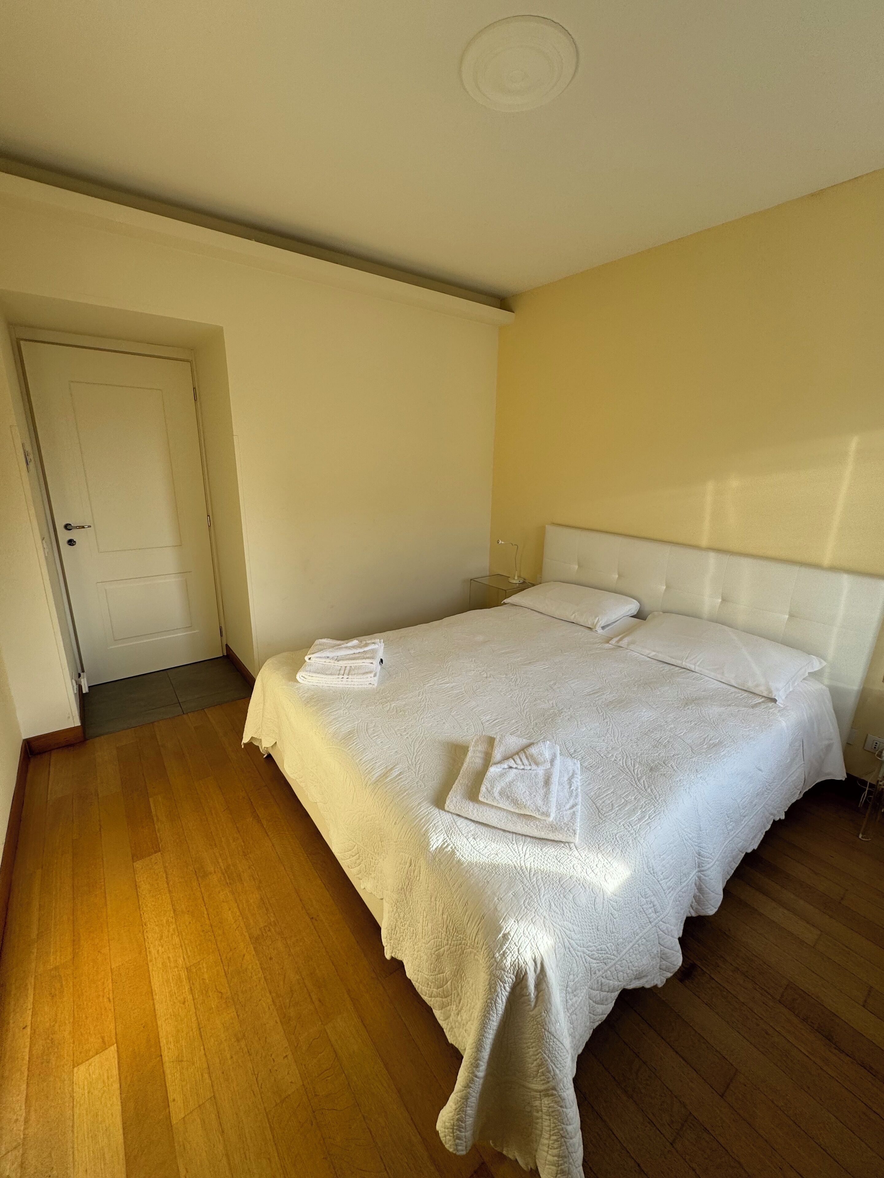 A neatly made bed with white linens and towels, a wooden floor, and a white door.