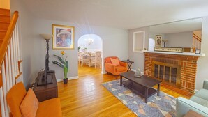 A living room with hardwood flooring, a fireplace, a dining area with a chandelier, and a staircase.