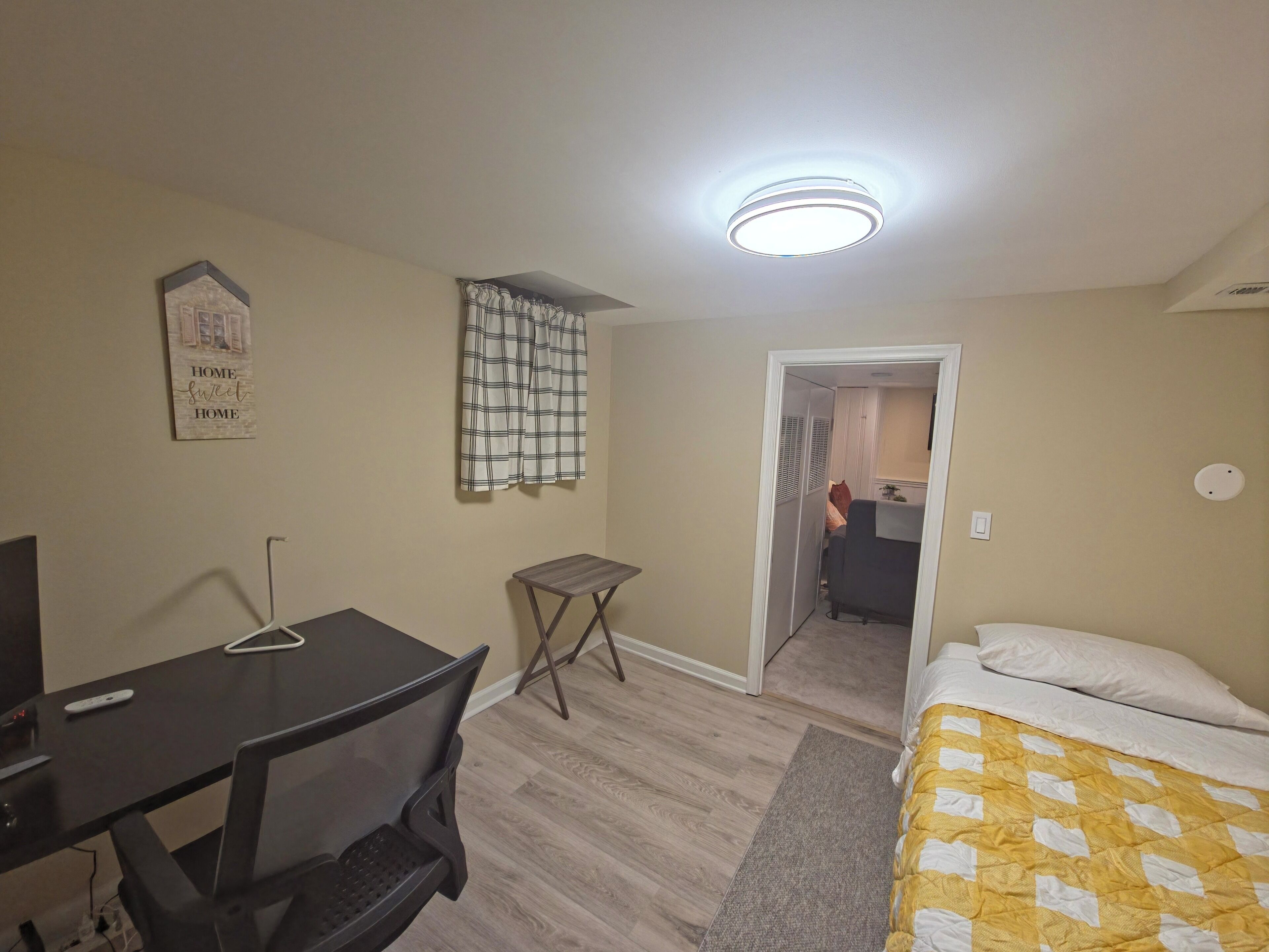 A small, well-lit room with a bed, a desk, and a chair. There is a window with a curtain, a wall-mounted light fixture, and a door leading to another room.