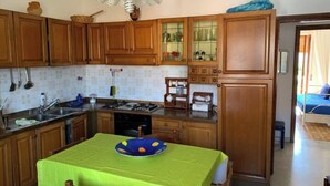 A compact kitchen with wooden cabinets, a dining table with a green tablecloth, and a small refrigerator. - 🏖️ Cozy Holiday Apartment 300m from the Sea - Policastro Bussentino #1 (Policastro Bussentino)