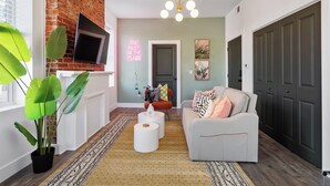 A modern living room with a brick accent wall, a flat-screen TV mounted above a fireplace, a white sofa with patterned cushions, and a large green plant.