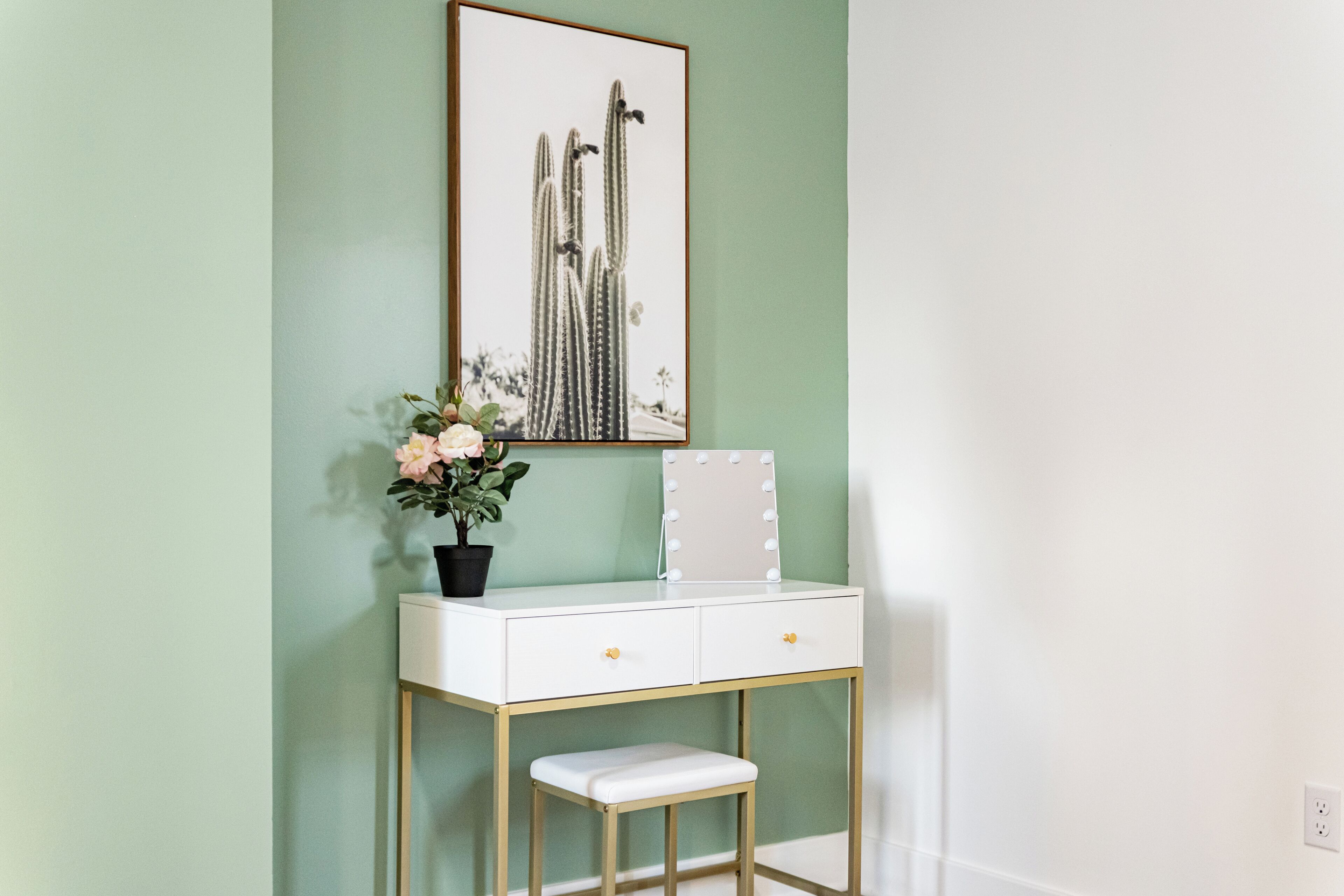 A white vanity with gold legs, a bench, and a framed cactus picture on the wall.