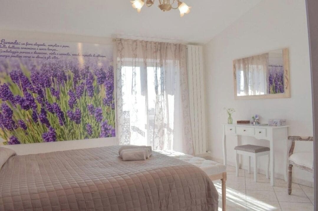 A bedroom with a large bed, a desk, and a chair. There is a window with sheer curtains and a painting of lavender flowers on the wall.