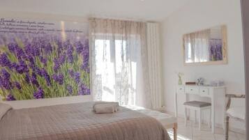 A bedroom with a large bed, a desk, and a chair. There is a window with sheer curtains and a painting of lavender flowers on the wall.