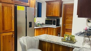A kitchen with wooden cabinets, a stainless steel refrigerator, a microwave, and two bar stools. - Luna by Suarez Properties | Jacuzzi, & BBQ (Santo Domingo Este)