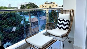 A balcony with a wicker chair and a small table, a chain-link fence, and buildings in the background. - Luna by Suarez Properties | Jacuzzi, & BBQ (Santo Domingo Este)