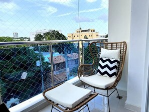 Uma varanda com cadeira de vime e uma pequena mesa, cerca de arame e edifícios ao fundo.
