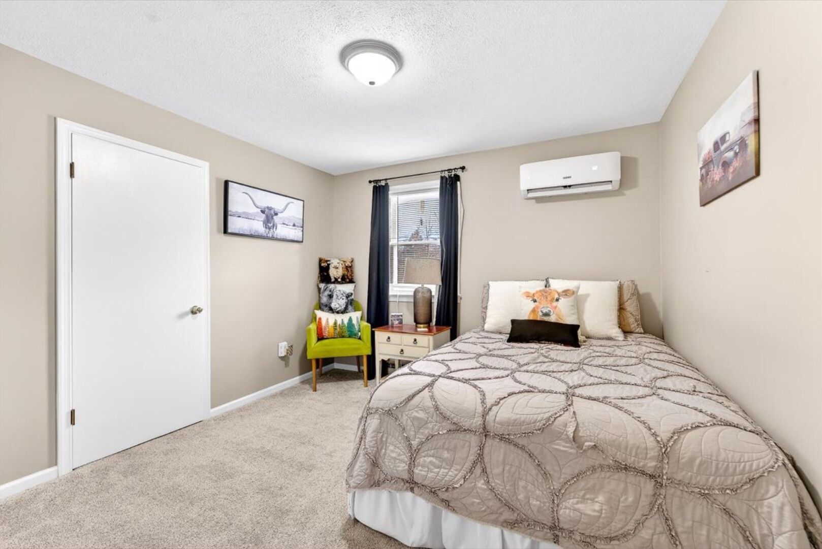 A bedroom with a bed, a chair, a nightstand, a window with curtains, and an air conditioning unit.
