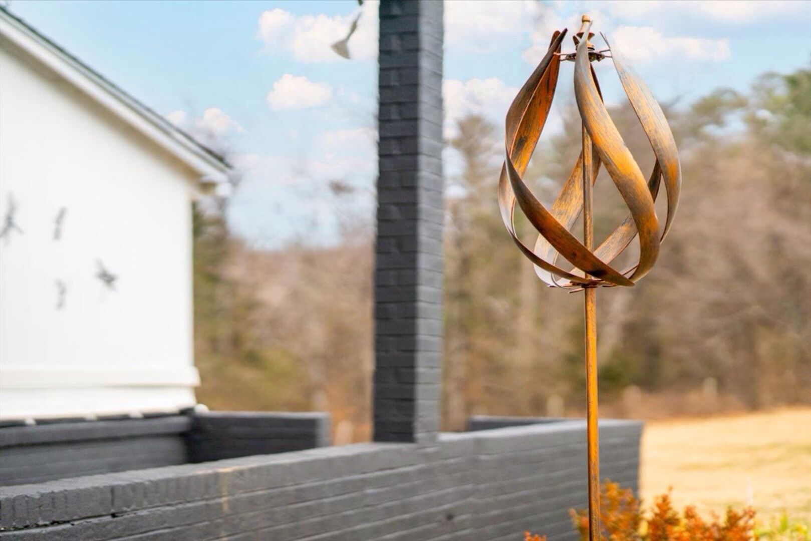 A metal wind chime hanging on a post in front of a building with a white exterior and a dark roof.