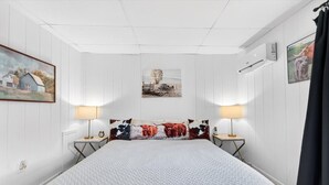 A bedroom with a large bed, two bedside tables with lamps, a wall-mounted air conditioner, and framed pictures on the walls.