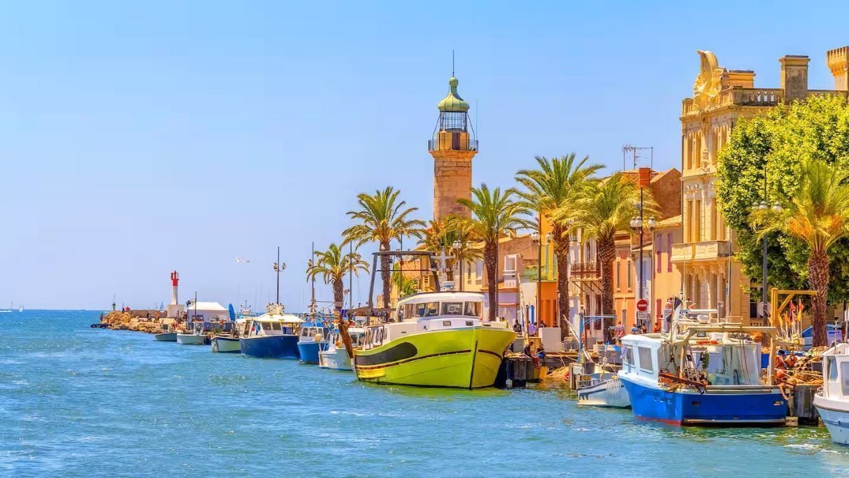 A waterfront with boats docked, a prominent lighthouse, and buildings with classical architecture.