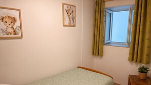 A small, single-bed room with a green patterned bedspread, a wooden headboard, a small bedside table with a plant, and two framed animal pictures on the wall.