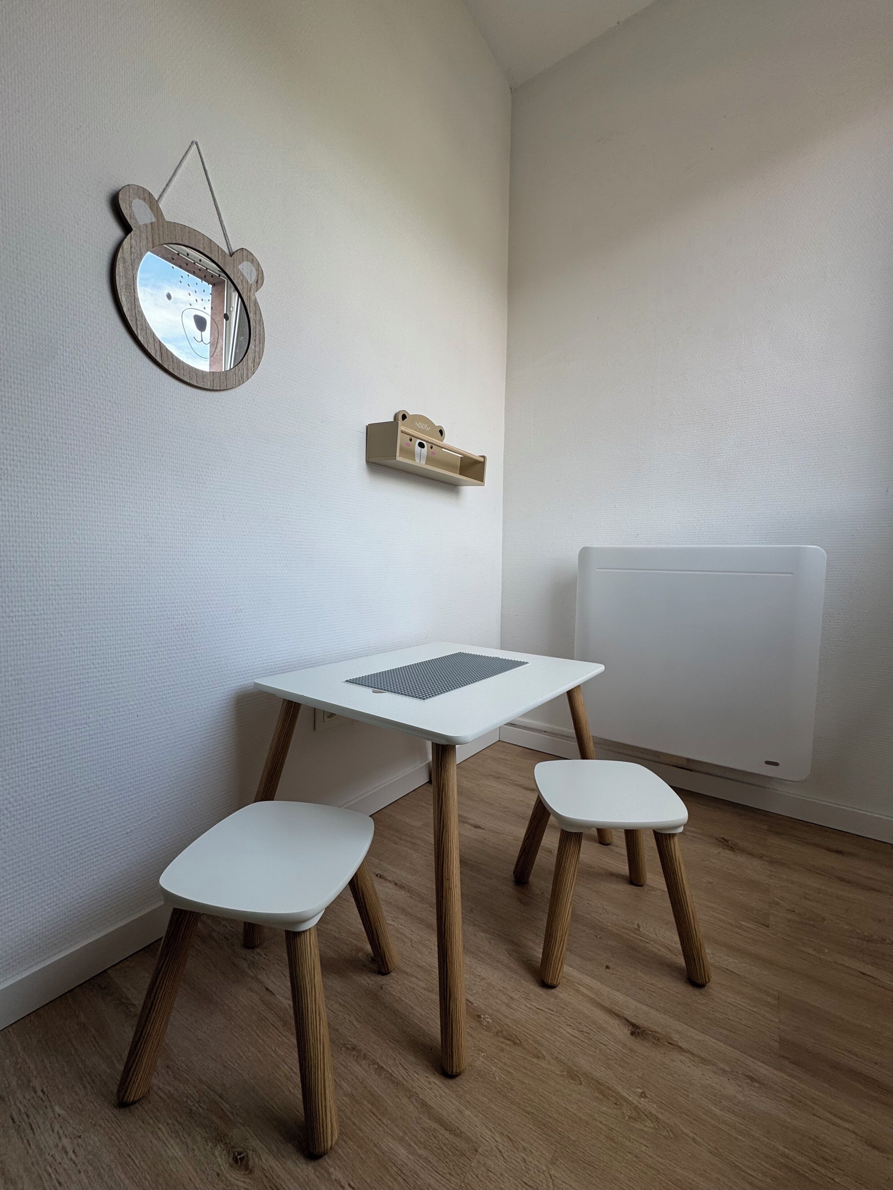 Une pièce avec une table blanche, deux chaises blanches, un miroir en bois en forme d’ours et un radiateur.